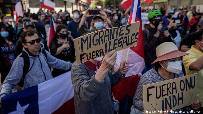 Imagen de personas en una manifestación contra la migración. 
Se muestran varias banderas chilenas.
Un cartel al centro de la imagen señala "Migrantes ilegales fuera", y otro tiene escrito el mensaje "Fuera Onu y INDH"